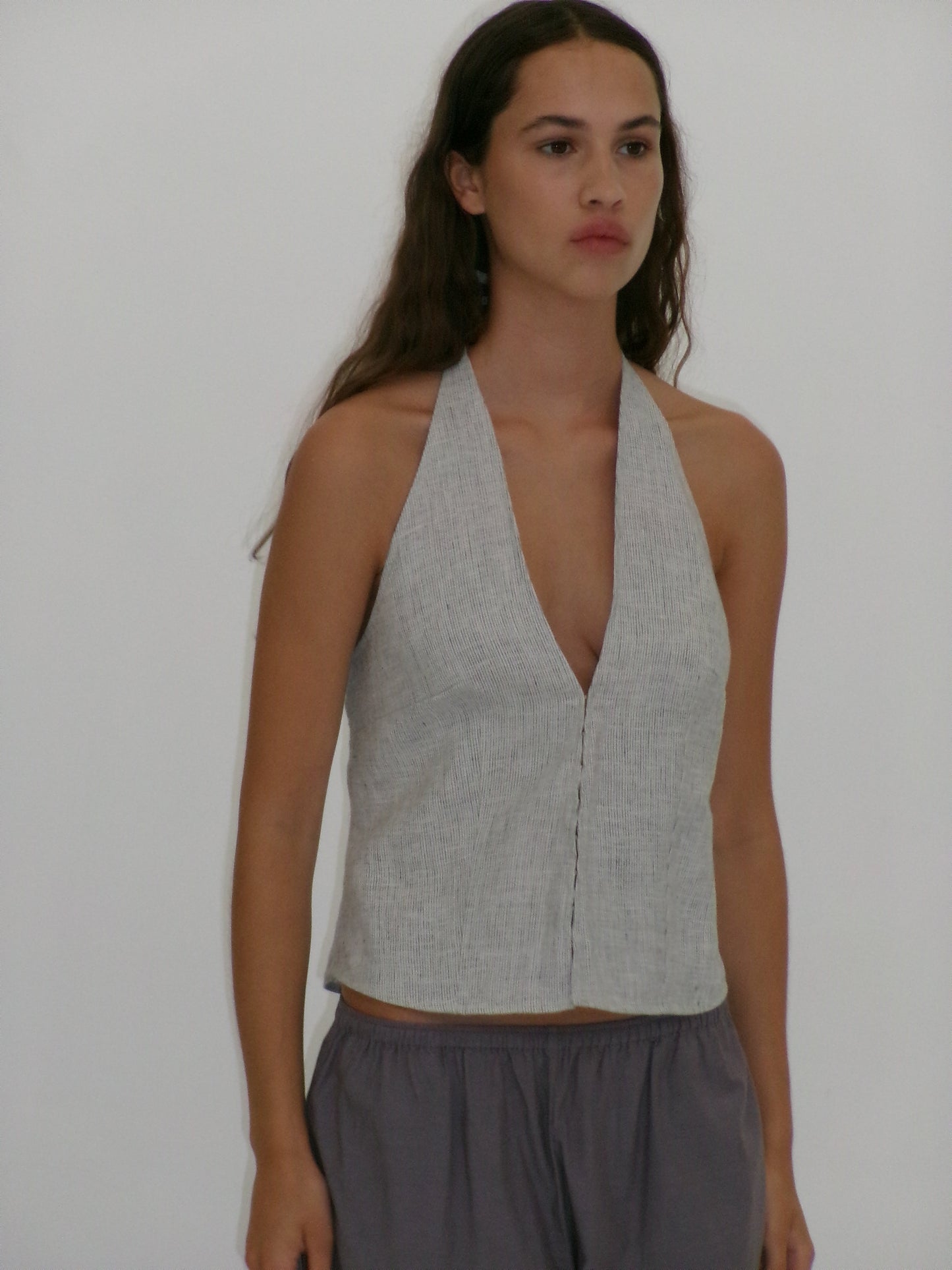 A woman with long brown hair wears the Close Halter Top - Fine Stripe and loose gray pants from Deiji Studios, standing against a plain white background.