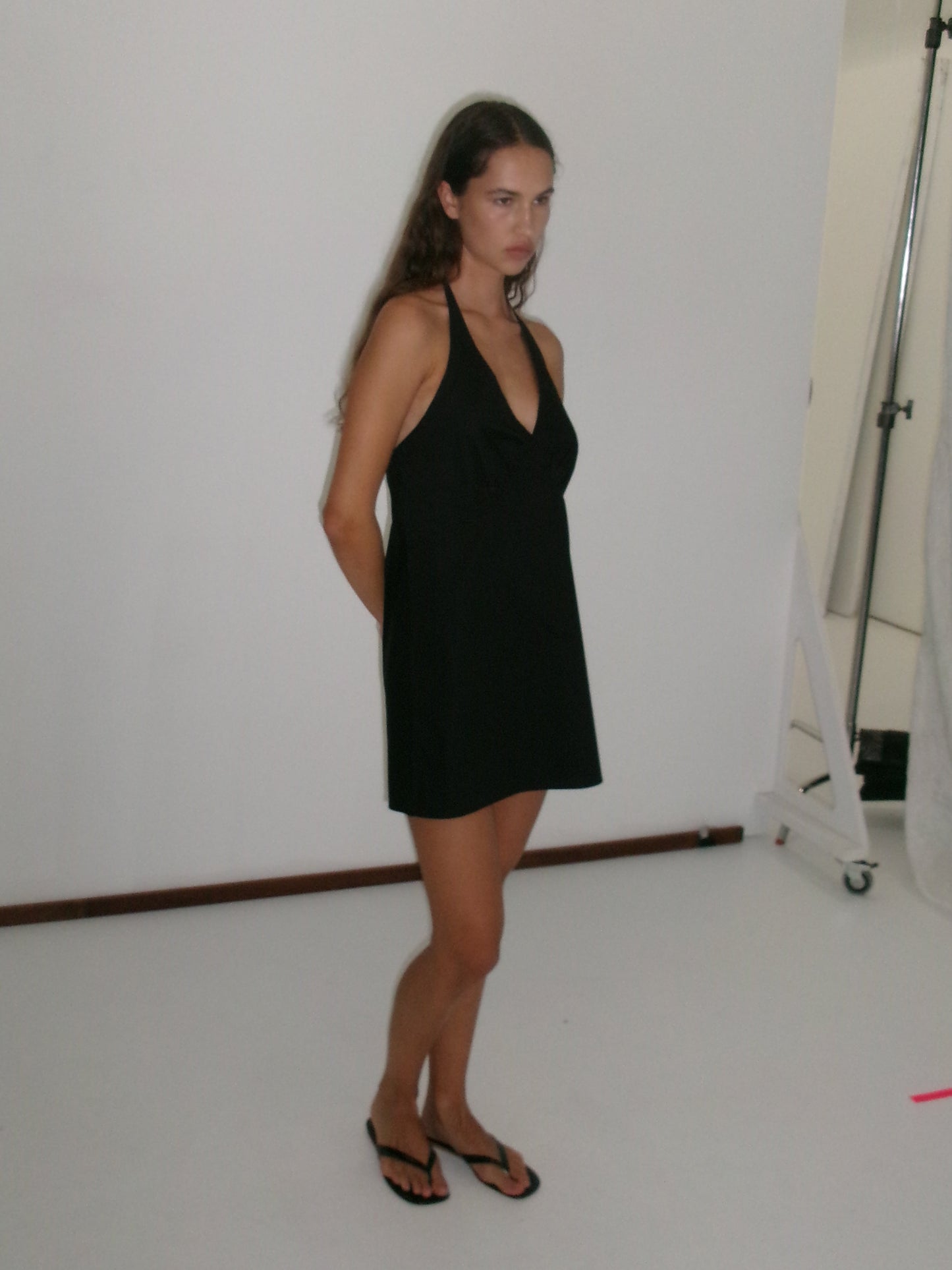 A woman with long brown hair wears the Cinched Halter Dress - Black, styled with black flip-flops, and stands with her hands behind her back in a white studio.