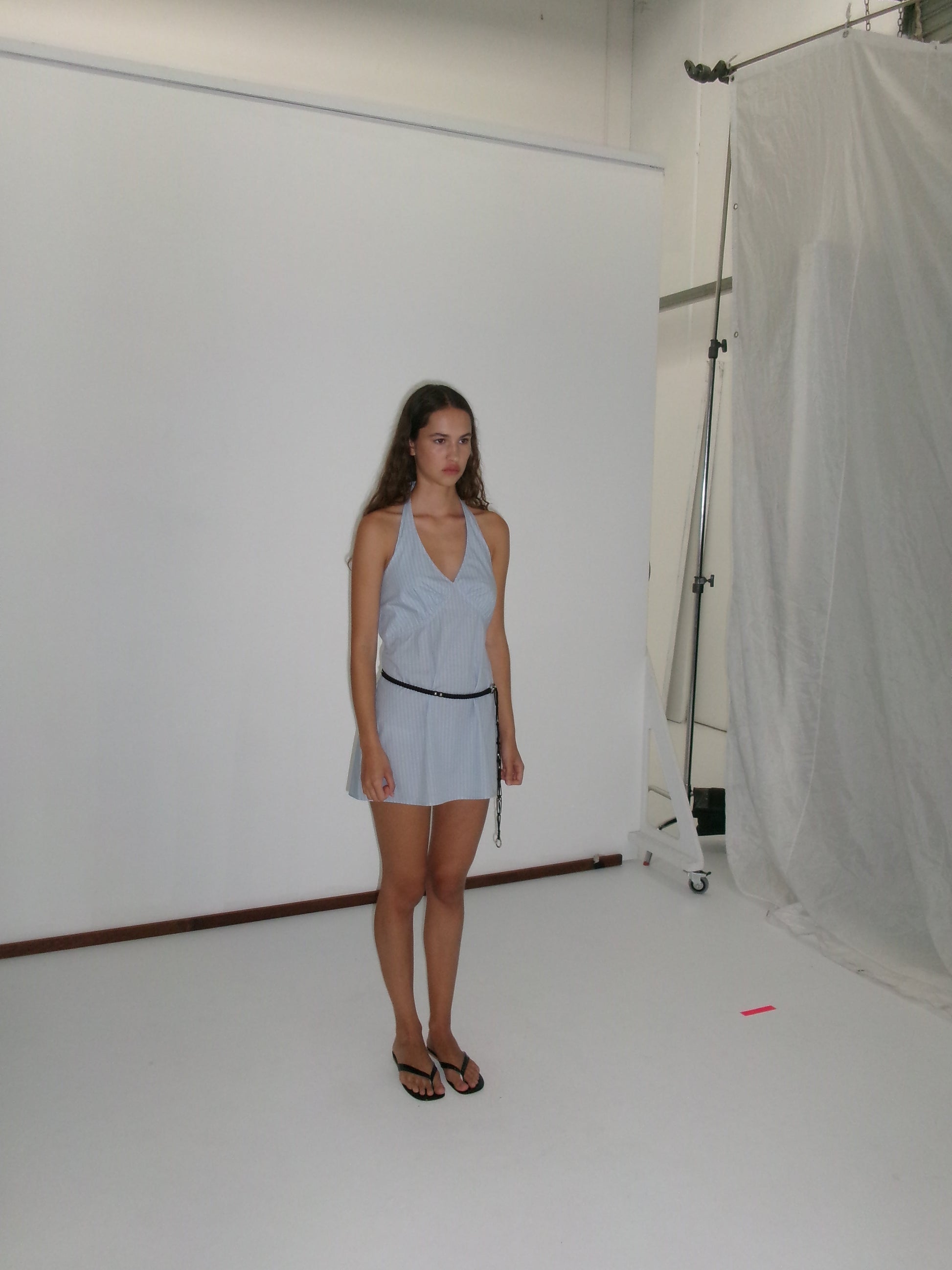 A young woman with long hair stands before a white backdrop, wearing the Cinched Halter Dress - Lake Blue Stripe by Deiji Studios, paired with black sandals and a belt. She gazes slightly to the side with a neutral expression.