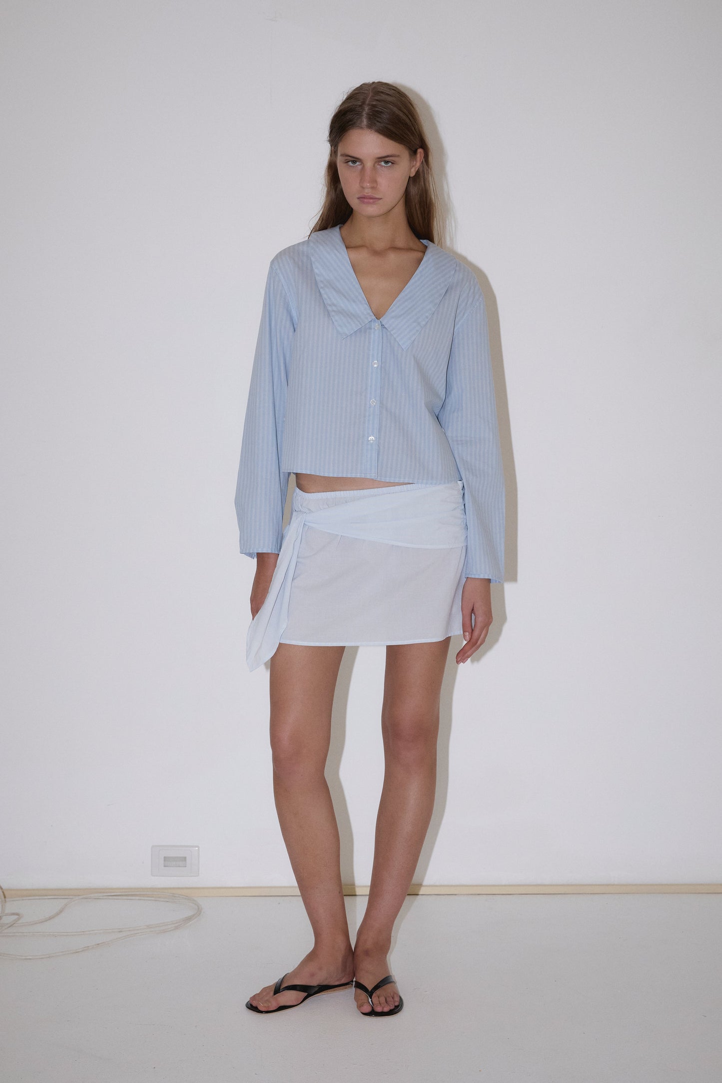 A woman with long straight hair stands against a plain white wall wearing the Deiji Studios Waist Button Shirt in Lake Blue Stripe, a white mini skirt with a sheer overlay, and black flip-flops.