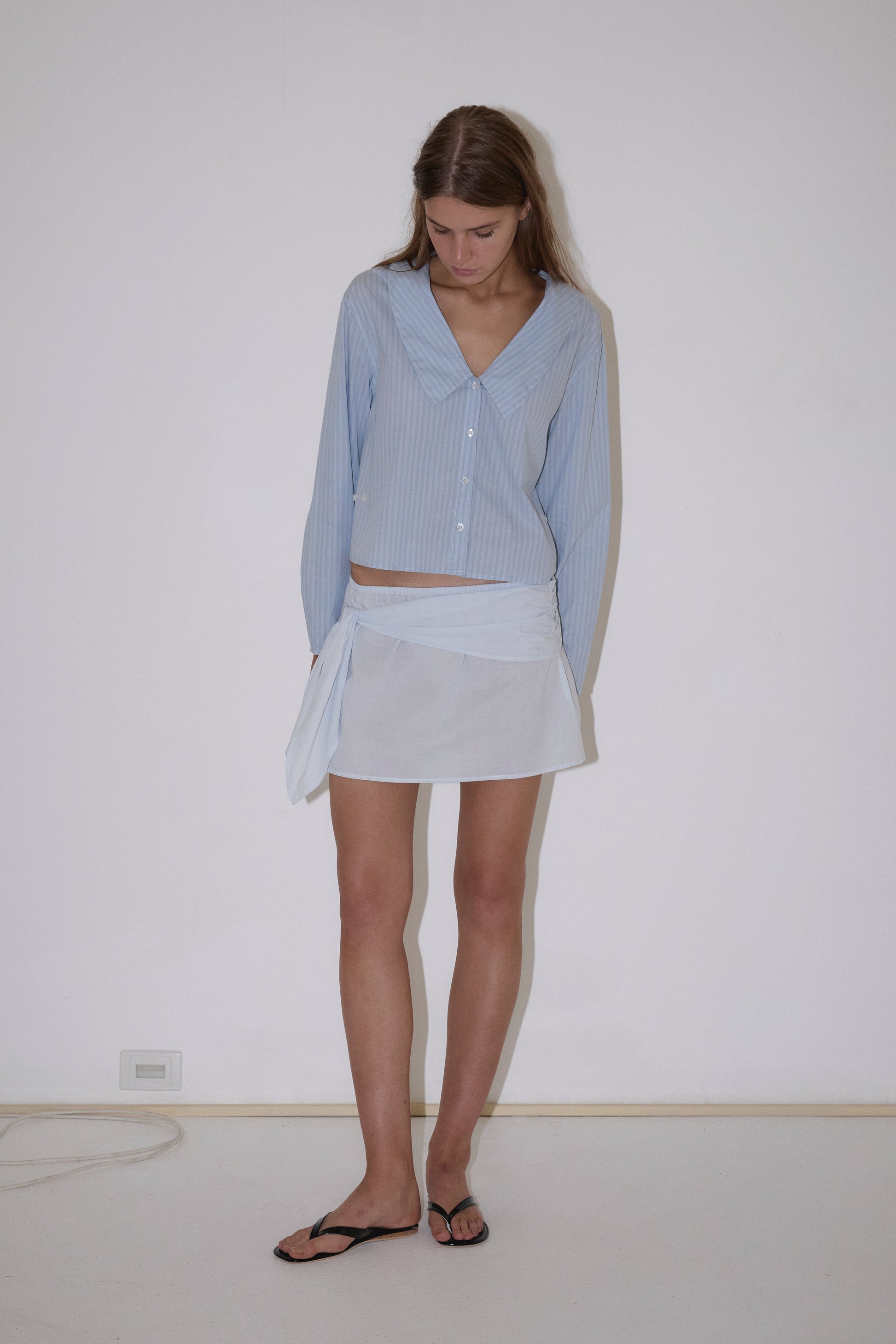 A woman stands indoors against a plain white wall, looking down. She wears a Deiji Studios Waist Button Shirt in Lake Blue Stripe, a white mini skirt with side tie, and black flip-flops.