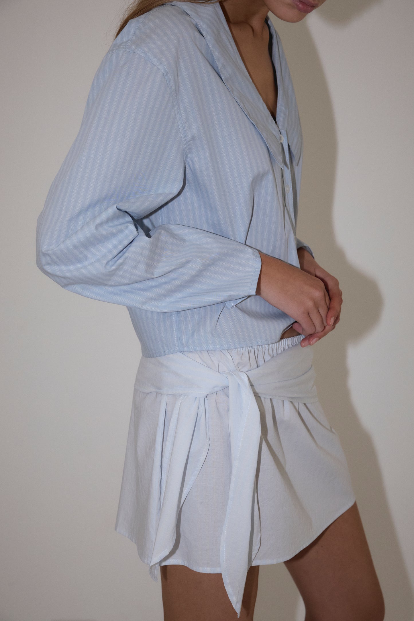 A woman wears the Deiji Studios Waist Button Shirt in Lake Blue Stripe, a lightweight collared blouse with subtle stripes, paired with a white waist-tied skirt. She stands against a plain light wall, face partially out of frame.