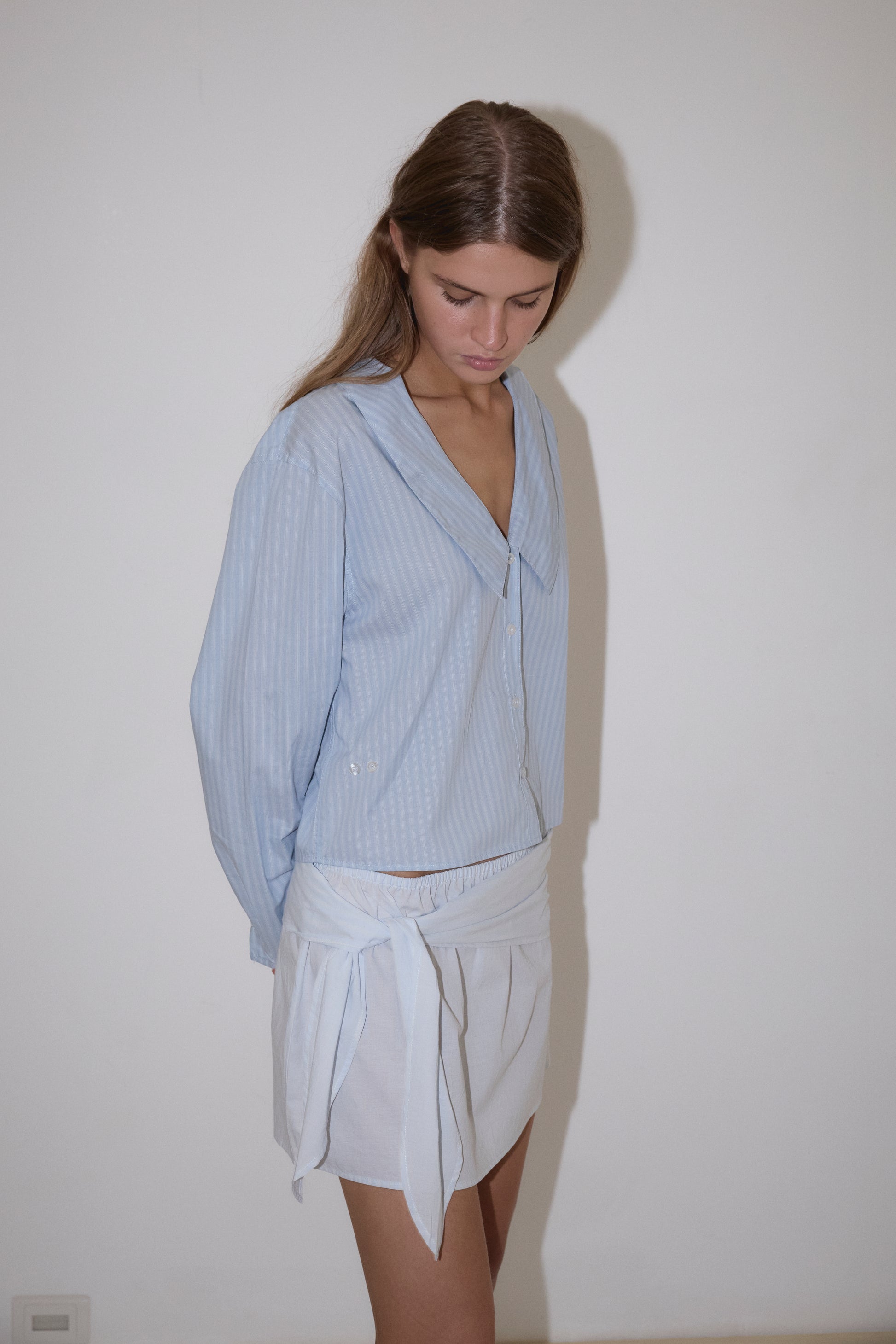 A woman with long brown hair stands against a white wall, looking down. She wears Deiji Studios' Waist Button Shirt in Lake Blue Stripe—a light blue, loose-fitting striped blouse—and a white wrap skirt, hands behind her back.