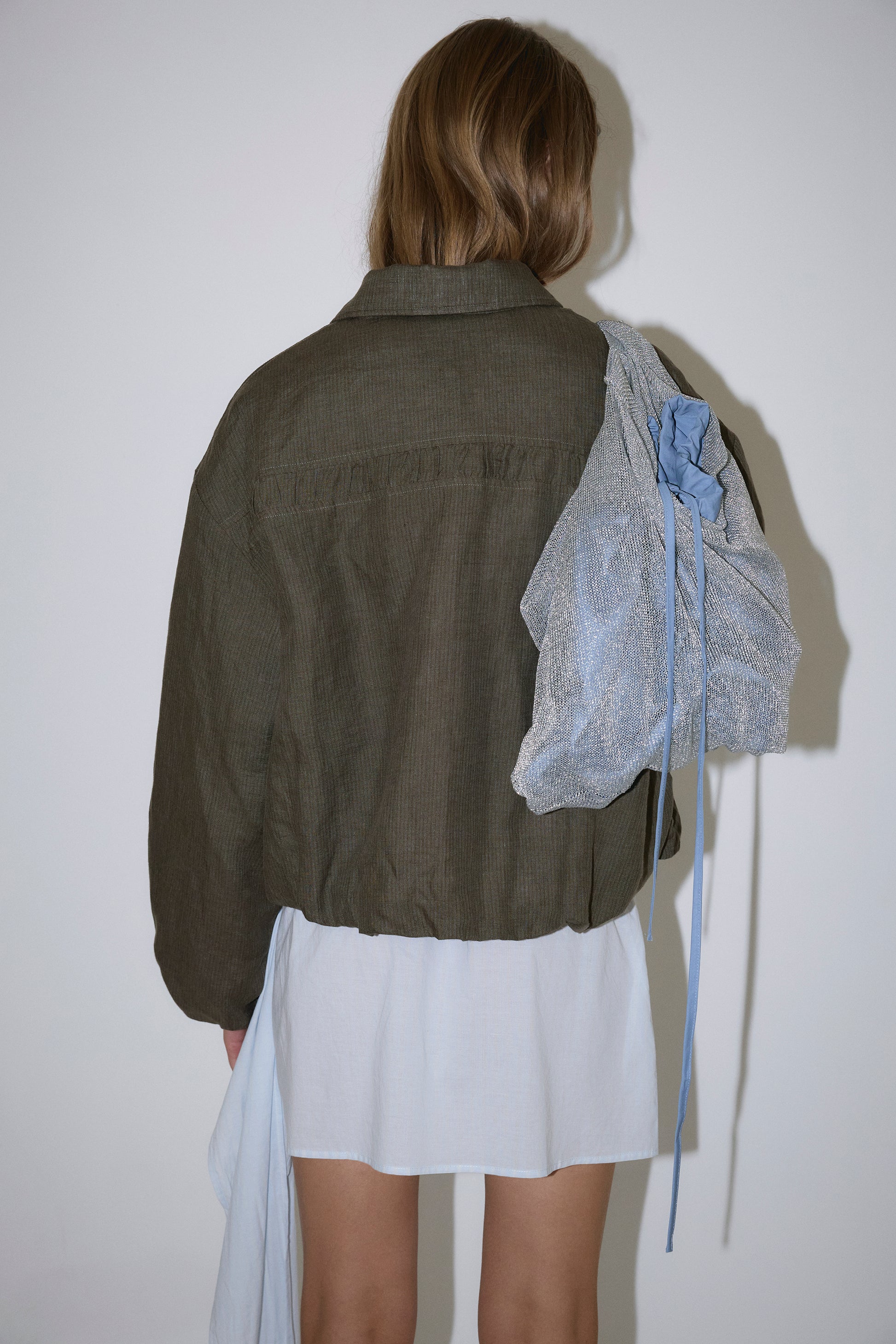 A person with long hair stands facing away, wearing the Pintuck Jacket - Khaki Stripe by Deiji Studios over a short white dress. They carry a textured gray bag with blue accents, set against a plain white background.