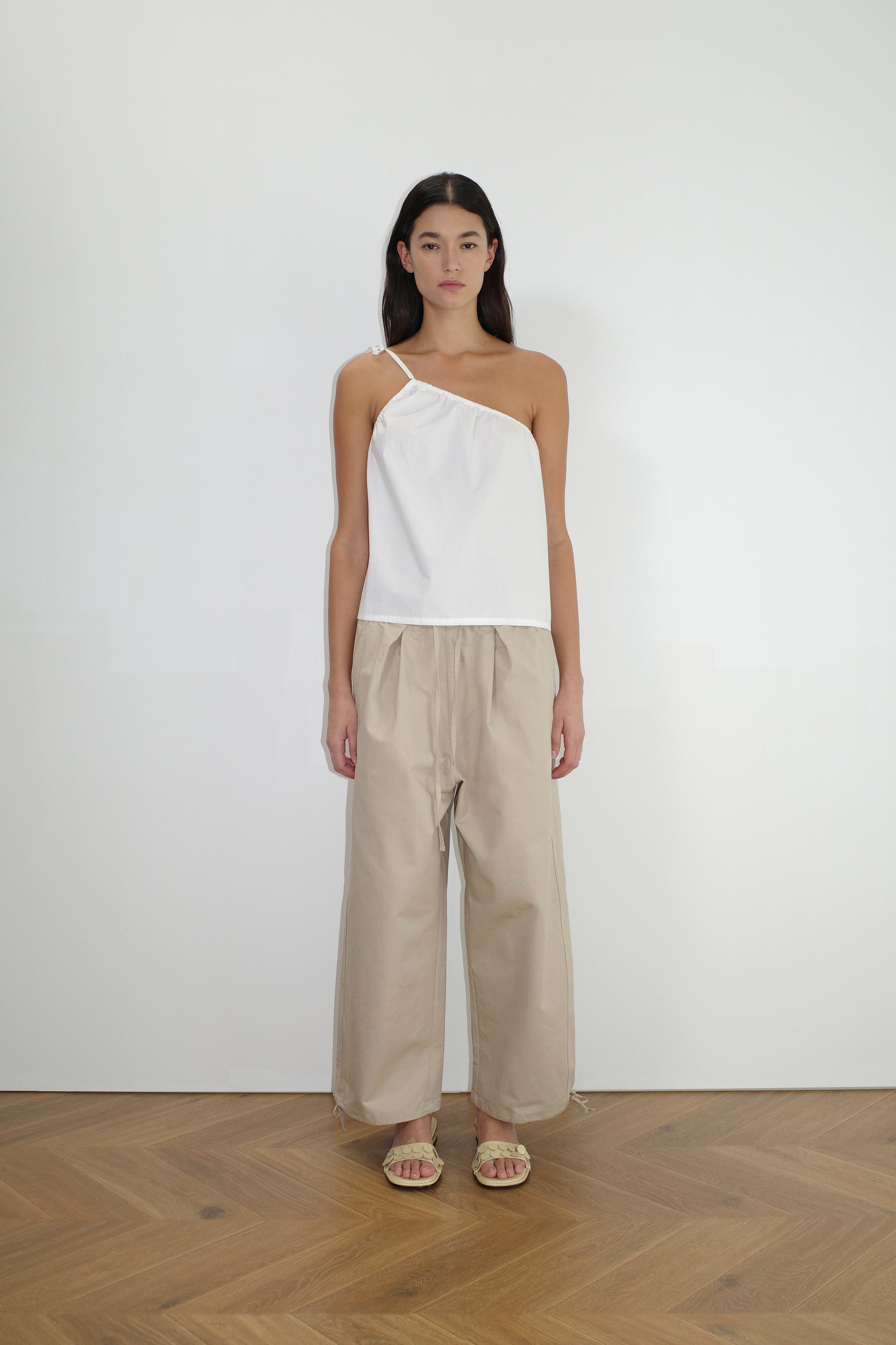 A woman stands indoors against a white wall, wearing a Deiji Studios white one-shoulder top, Drawcord Wide Leg Pant in cream, and sandals. She has long dark hair and a neutral expression, standing on a wooden floor.
