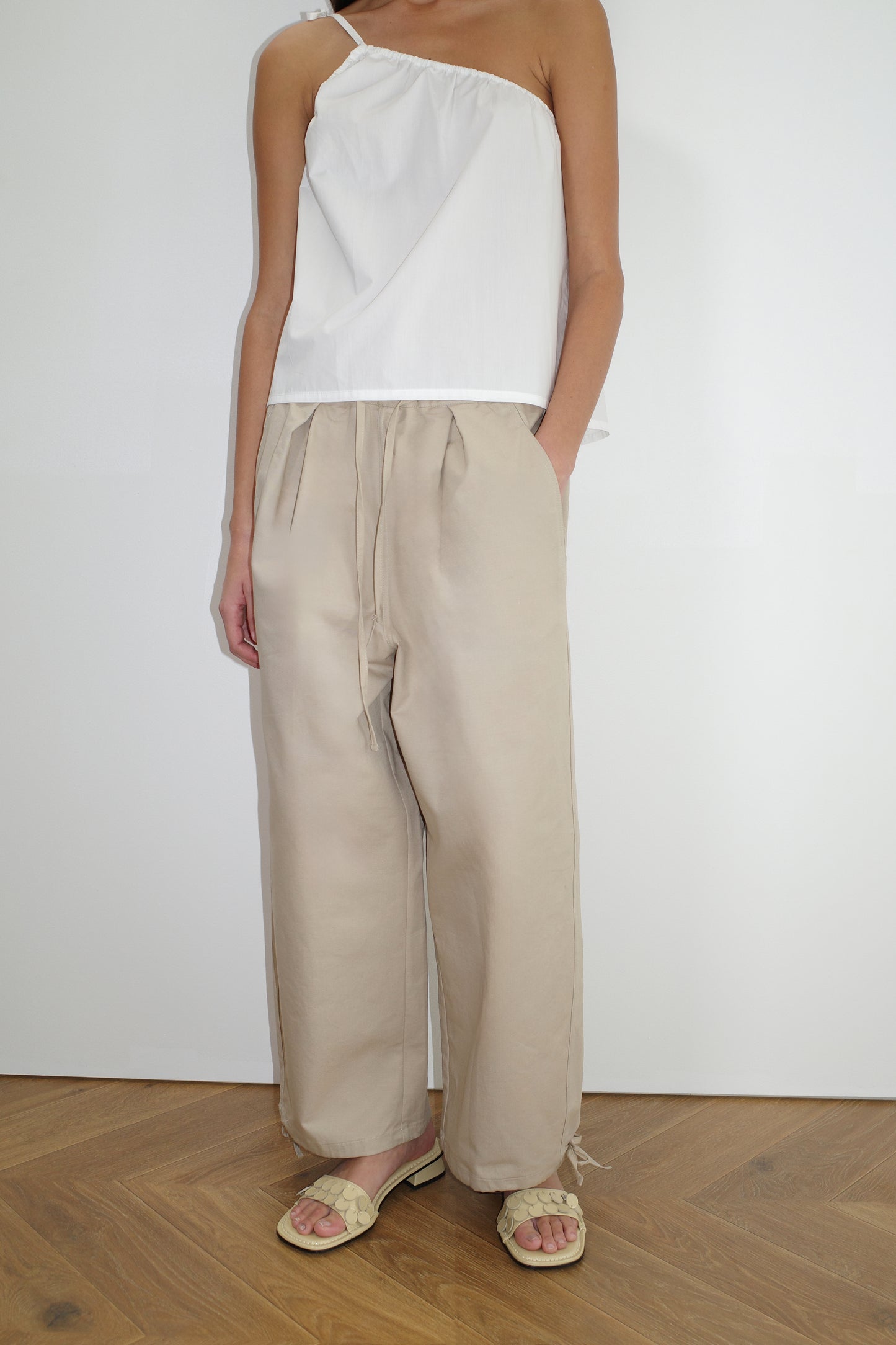 A person in a white one-shoulder top, the Drawcord Wide Leg Pant - Cream, and light-colored sandals stands on a wooden floor against a plain white wall. Their head is not visible in the image.
