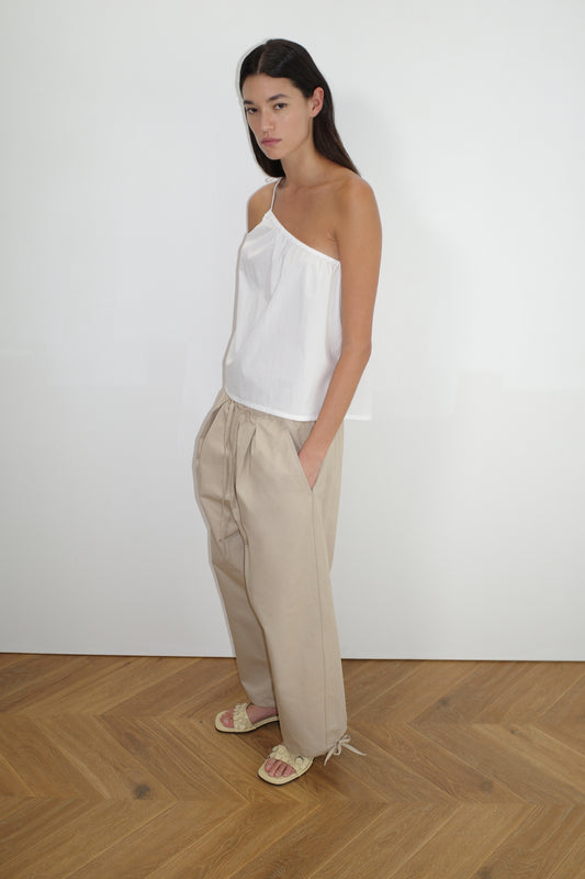 A woman with long dark hair stands indoors against a white wall, wearing a Deiji Studios one-shoulder top and the Drawcord Wide Leg Pant in Cream, paired with light sandals on a classic herringbone wooden floor.