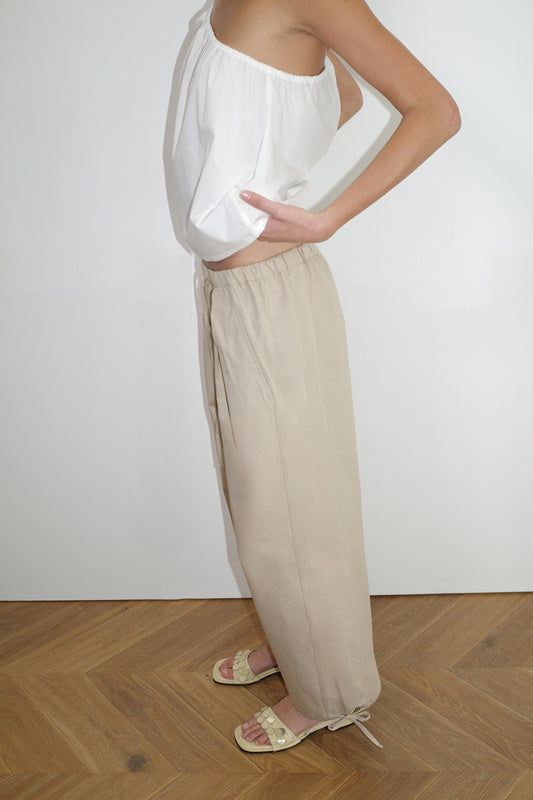 A person stands sideways against a white wall, wearing a Deiji Studios white off-shoulder top, Drawcord Wide Leg Pant in cream, and cream-colored sandals with ankle ties on a herringbone wooden floor.