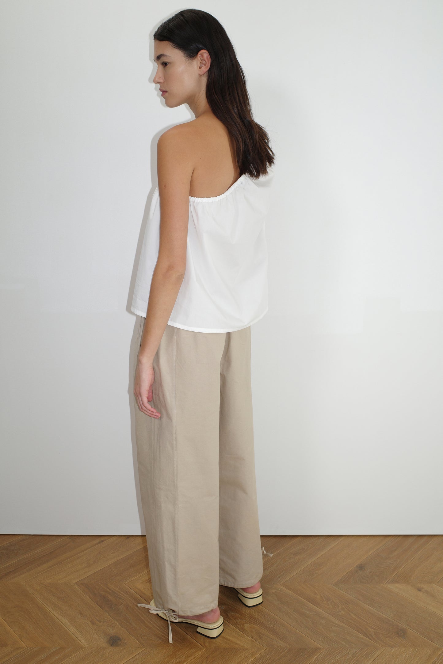 A woman stands sideways against a white wall, wearing a white off-shoulder sleeveless top and Deiji Studios Drawcord Wide Leg Pant in Cream with flat shoes on a wooden floor.