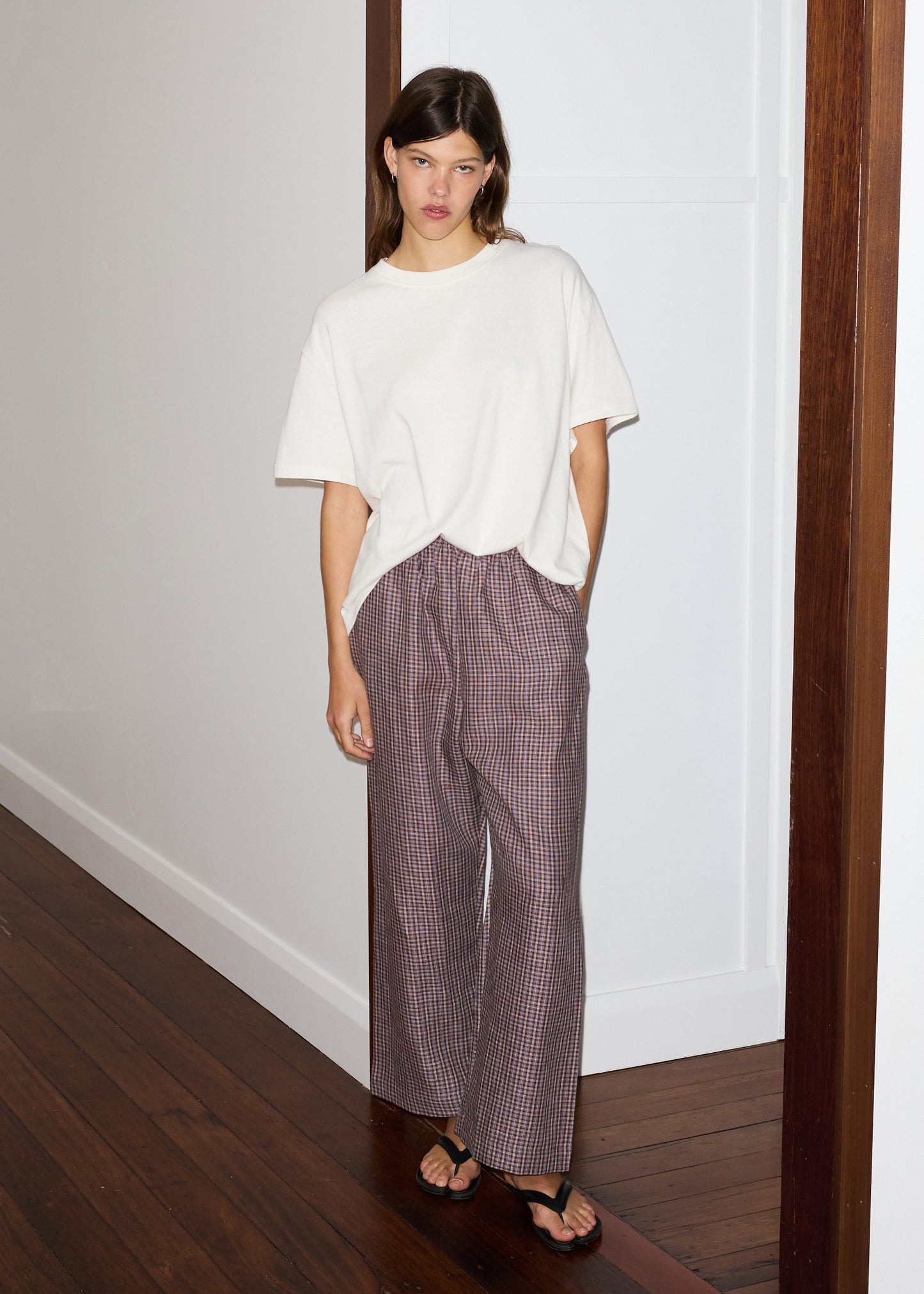 A woman stands indoors by a white wall, wearing a loose white t-shirt, linen ease trouser - russet check from Deiji Studios in wide-leg plaid, and black sandals. One hand is in her pocket as she looks relaxed on the dark wood floor.