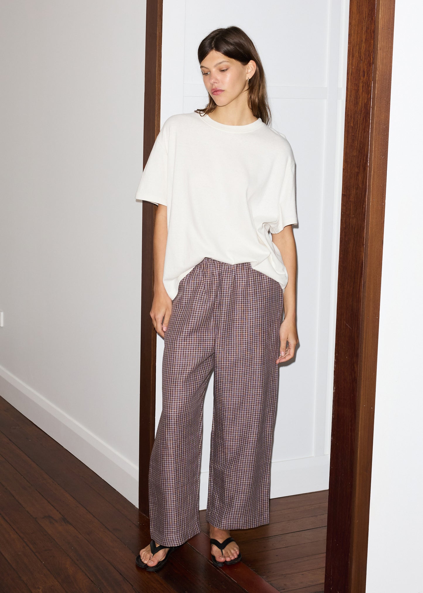 A woman stands indoors wearing a loose white tee, linen ease trouser - russet check, and black flip-flops. She has straight brown hair, one hand in her pocket, leaning against a white wall with wooden trim and floors.