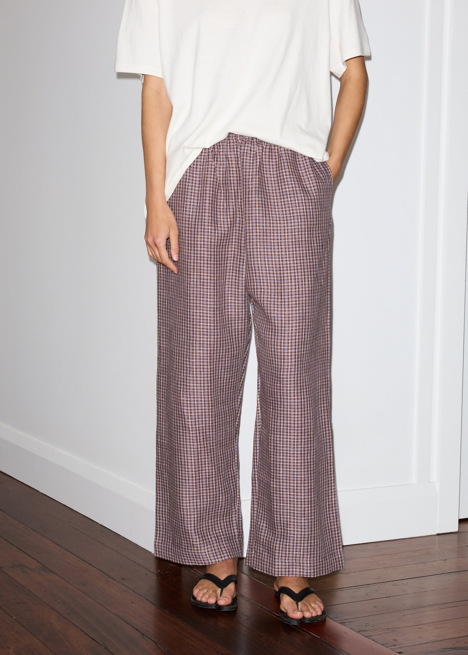 A person stands indoors wearing Deiji Studios' linen ease trouser - russet check, featuring a wide-leg plaid design, paired with a loose white t-shirt and black sandals. Their left hand is in their pocket; a wooden floor contrasts the white wall background.