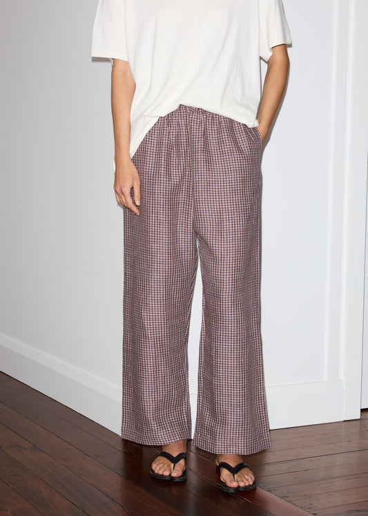 A person stands indoors wearing Deiji Studios' linen ease trouser - russet check, featuring a wide-leg plaid design, paired with a loose white t-shirt and black sandals. Their left hand is in their pocket; a wooden floor contrasts the white wall background.