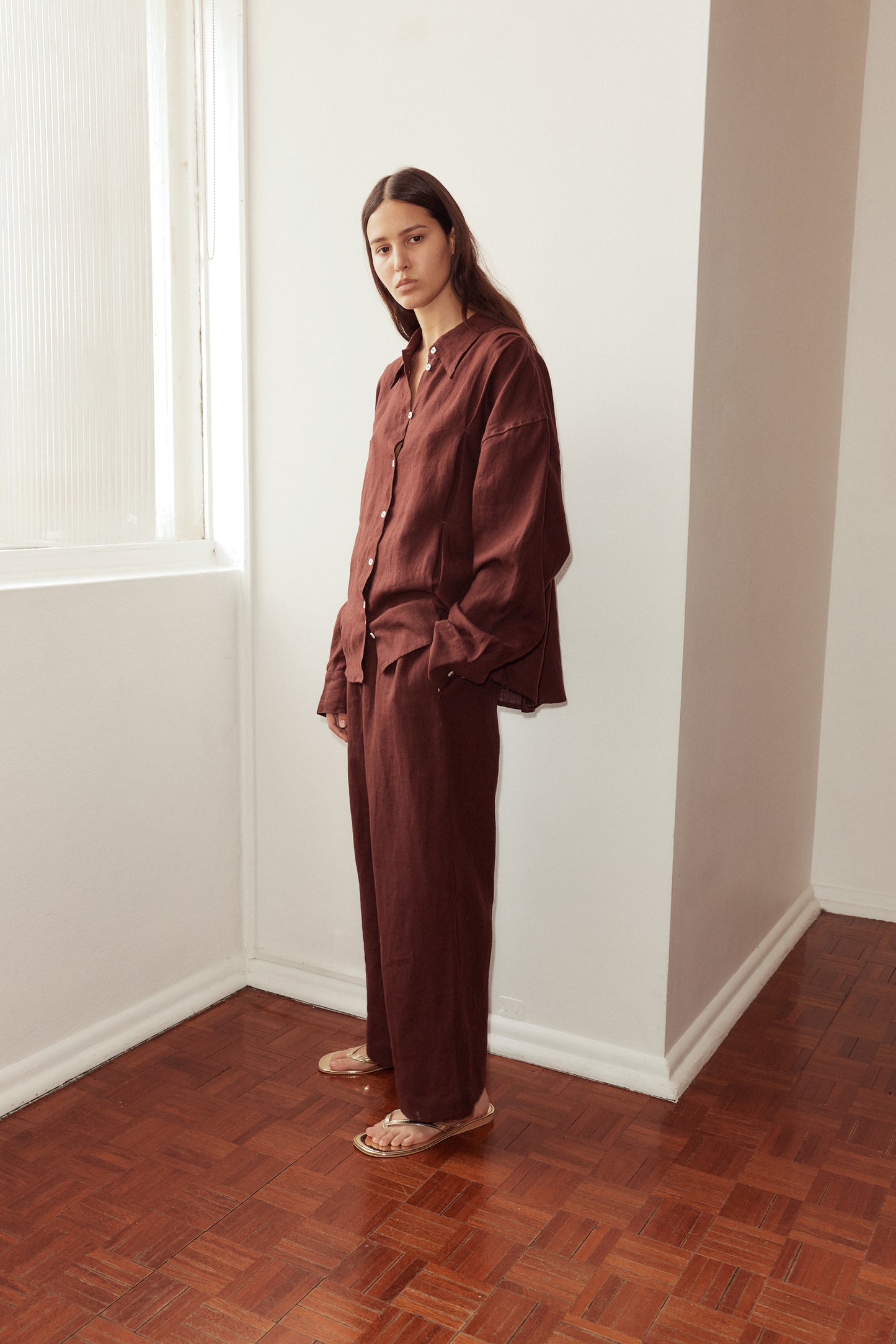 Female model standing against a window wearing the Tack Set by Deiji Studios in burgundy. Relaxed shirt features dropped shoulder, raglan sleeve and button front, pairs back with relaxed full length pant.