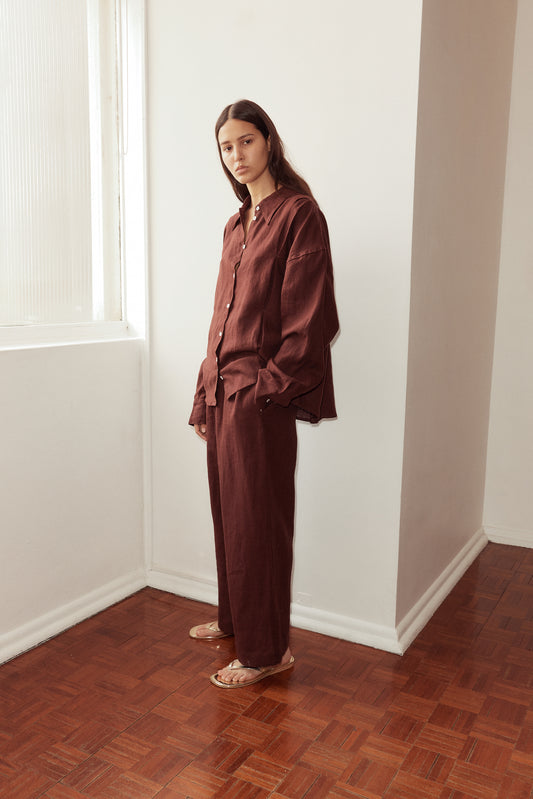 Female model standing against a window wearing the Tack Set by Deiji Studios in burgundy. Relaxed shirt features dropped shoulder, raglan sleeve and button front, pairs back with relaxed full length pant.