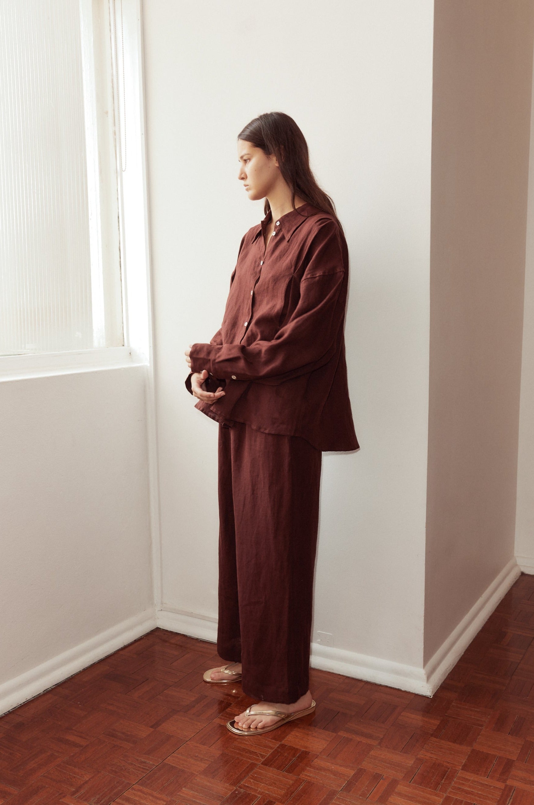 Female model standing against a window wearing the Tack Set by Deiji Studios in burgundy. Relaxed shirt features dropped shoulder, raglan sleeve and button front, pairs back with relaxed full length pant, styled with gold flip flops.