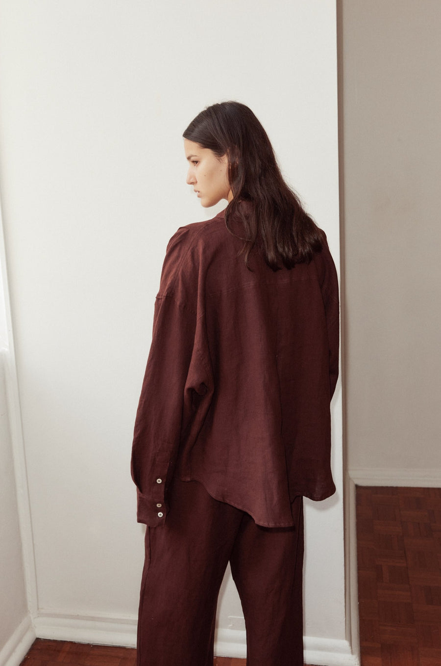 Back shot of female model wearing the Tack Set by Deiji Studios in burgundy. Relaxed shirt features dropped shoulder, dropped back yoke, raglan sleeve and button cuff.