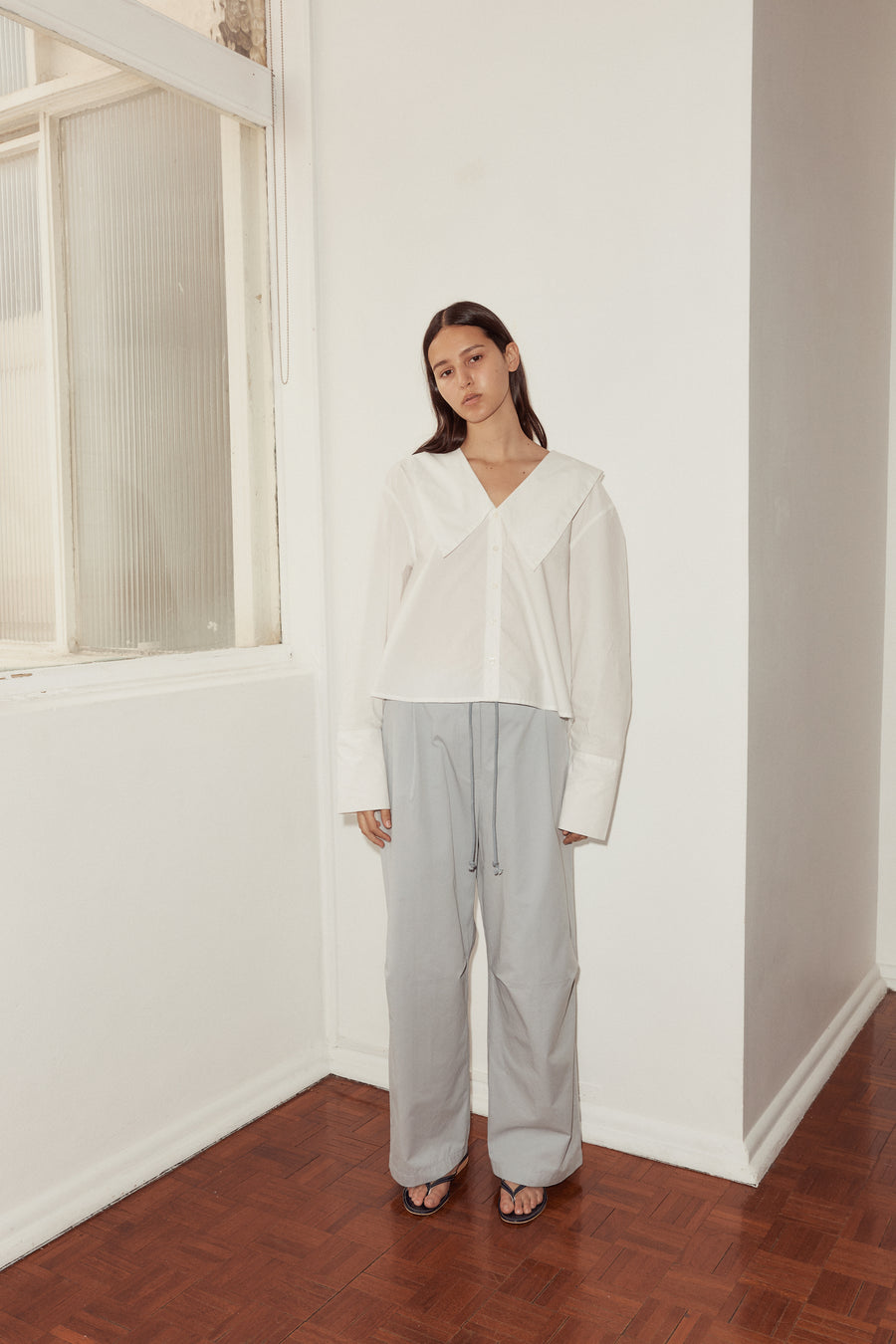 Long shot of female model wearing Oversized Collared Shirt in white, styled with sky blue Bartack Pants and black flip flops. Model stands in the corner of a white wall with window to the left.