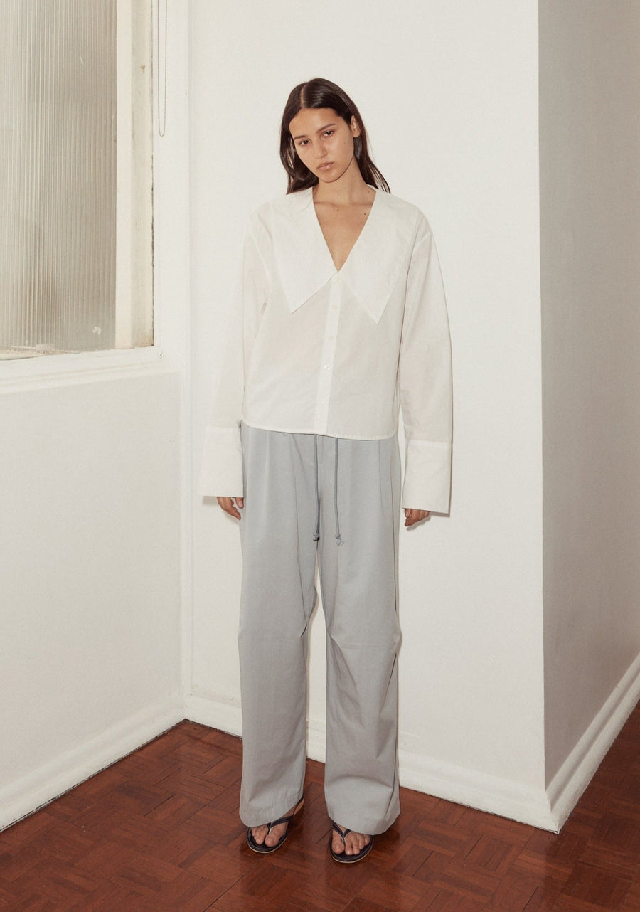 mid shot of female model wearing Deiji Studios sky blue cotton twill pants, with a relaxed fit, tucks at the knee and drawcord waist. Pant is styled with a Deiji Studios white shirt featuring an oversized collar, button front and wide cuff