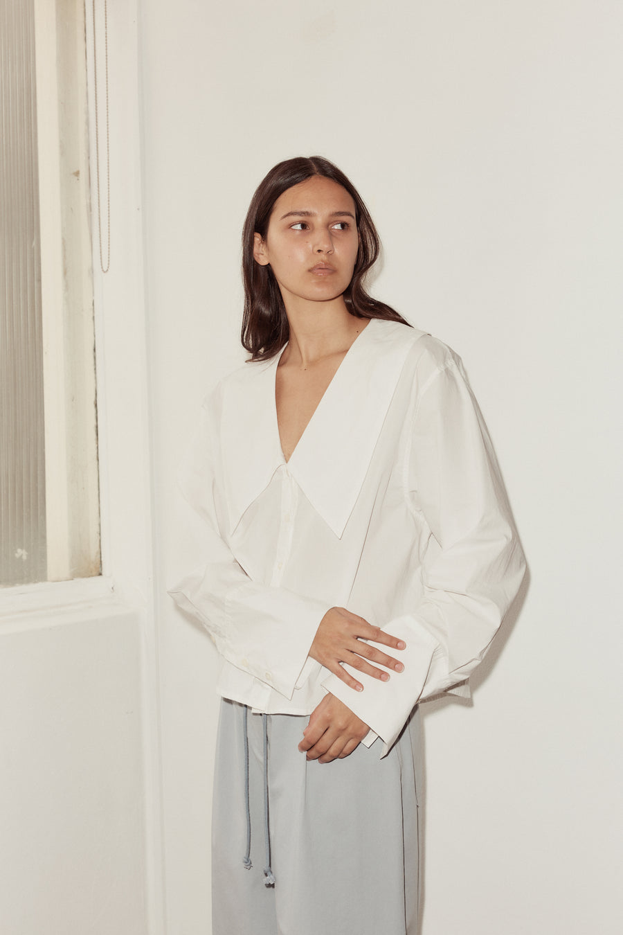 Mid shot of female model wearing Oversized Collared Shirt in white, model looks to the side with one hand touching the cuff of the other hand.