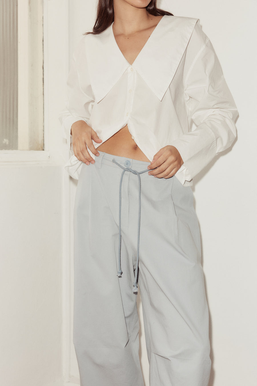 close up shot of female model wearing Deiji Studios sky blue cotton twill pants, features a drawcord waist, belt loops, and button closure at waist. Model wears Deiji Studios white oversized collared shirt.