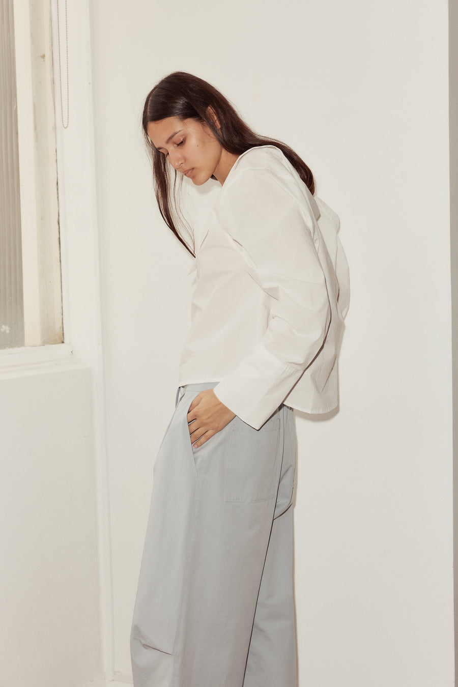 side shot of female model putting hand in the side pocket of Deiji Studios sky blue cotton twill pants. Pant has deep side and back pockets, and a tuck at each knee.