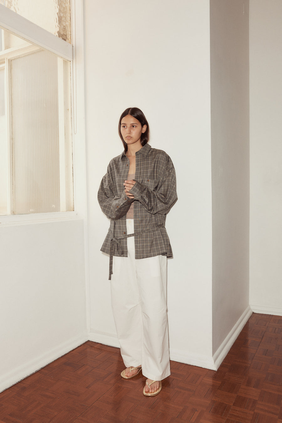Mid shot of female model standing near a window wearing the Wool Shirt in Everyday Check by Deiji Studios, styled with the Bartack Pant in white. Shirt features pointed collar, button front with single breast pocket and self fabric waist tie in subtly lustrous wool check.