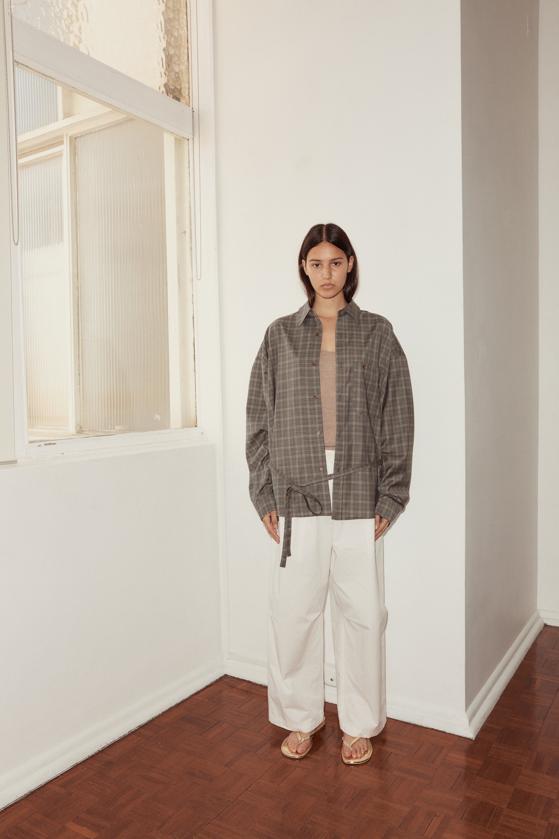 Mid shot of female model wearing the Wool Shirt in Everyday Check by Deiji Studios, styled with the Bartack Pant in white. Shirt features pointed collar, button front with single breast pocket and self fabric waist tie in subtly lustrous wool check.