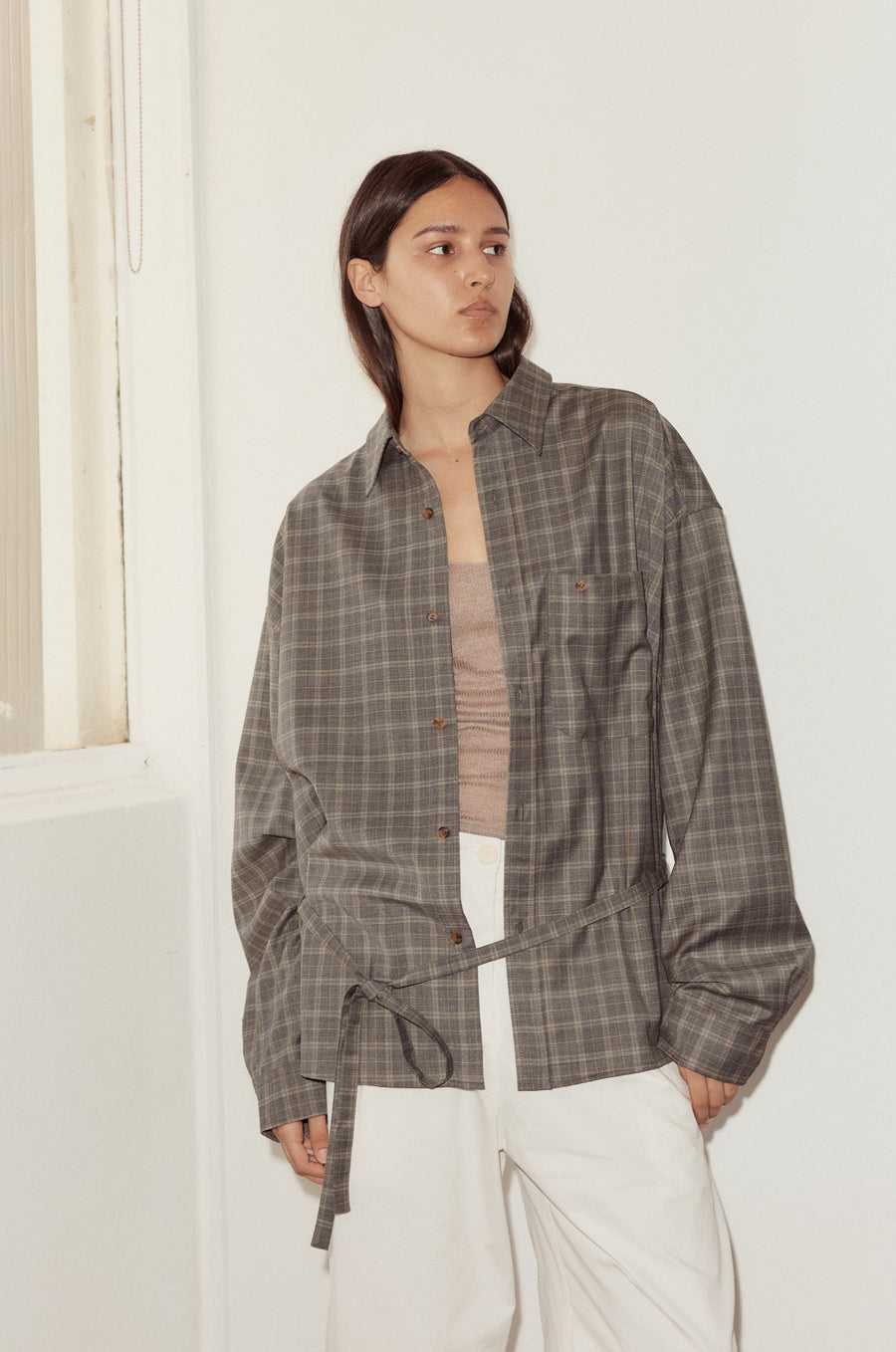 Mid shot of female model wearing the Wool Shirt in Everyday Check by Deiji Studios, styled with the Bartack Pant in white. Shirt features pointed collar, button front with single breast pocket and self fabric waist tie in subtly lustrous wool check.