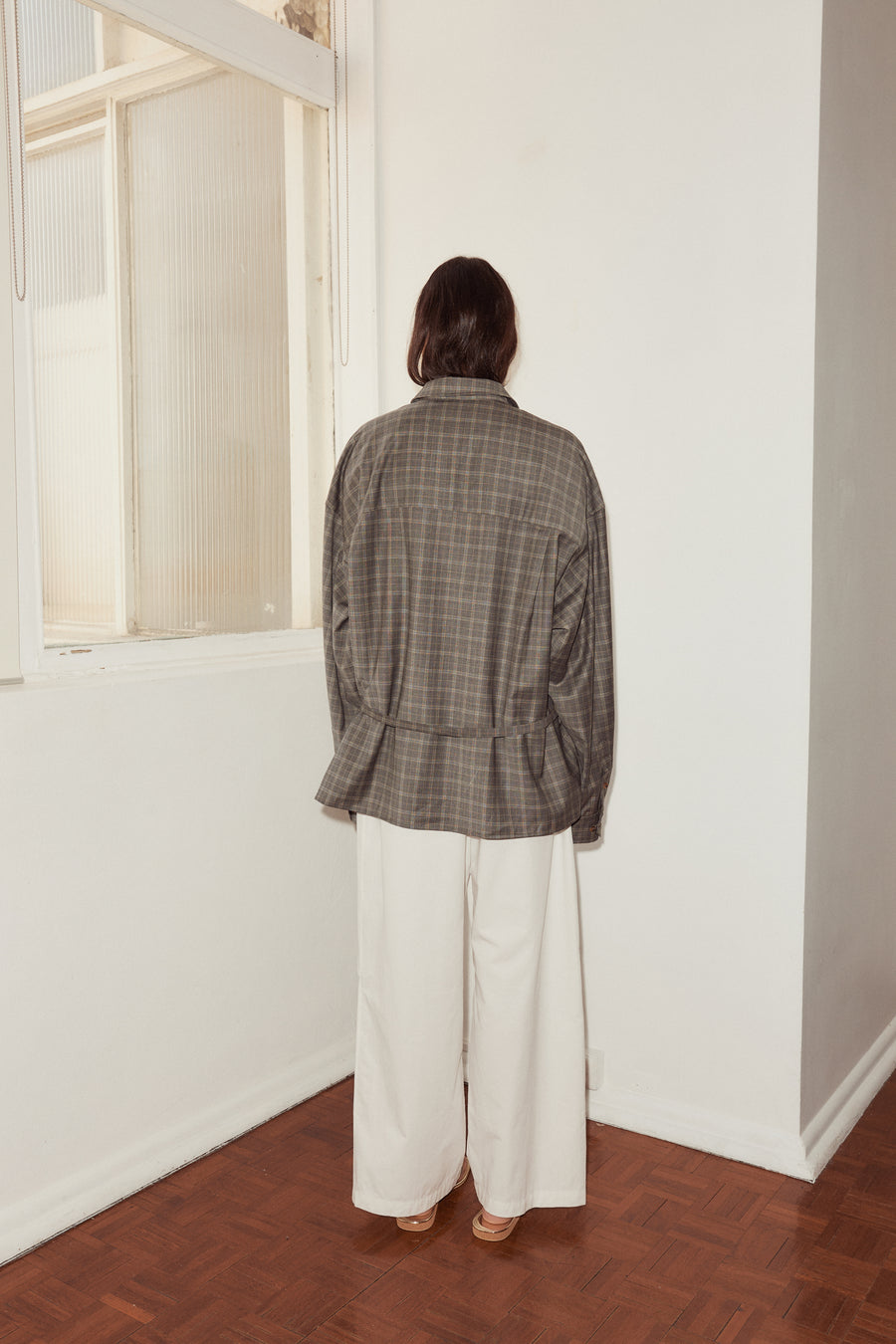 Back shot of female model wearing the Wool Shirt in Everyday Check by Deiji Studios, styled with the Bartack Pant in white. Shirt features dropped shoulder with dropped back yoke for a relaxed fit and self fabric waist tie.