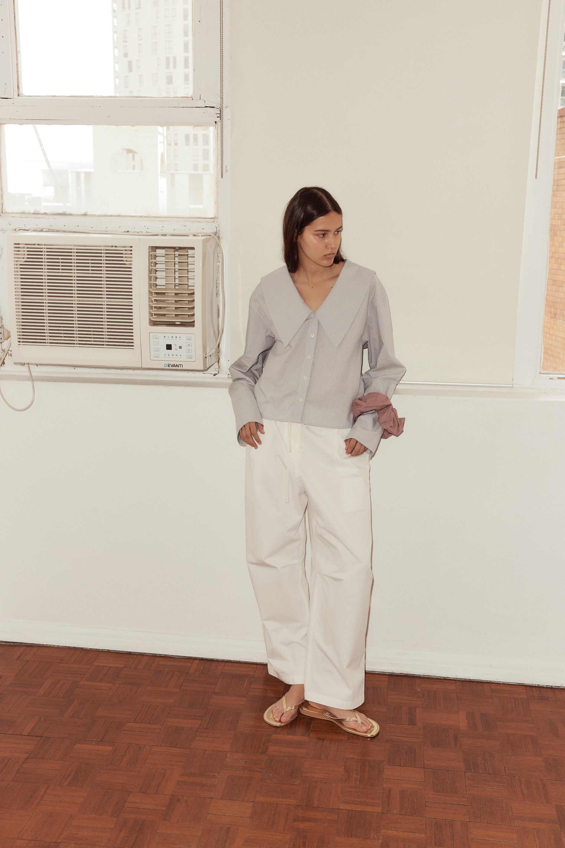 Long shot of female model wearing the Oversized Collared Shirt in Dream Stripe, Bartack Pants in White. Styled with gold flip flops and scrunchie worn on wrist. Model stands in from of an old air conditioner lodged in the wall.