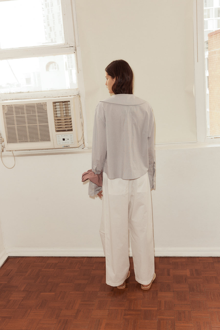 Back shot of female model wearing the Oversized Collared Shirt in Dream Stripe, Bartack Pants in White. Collar flips down over shoulders in a soft curve.Model stands in from of an old air conditioner lodged in the wall.