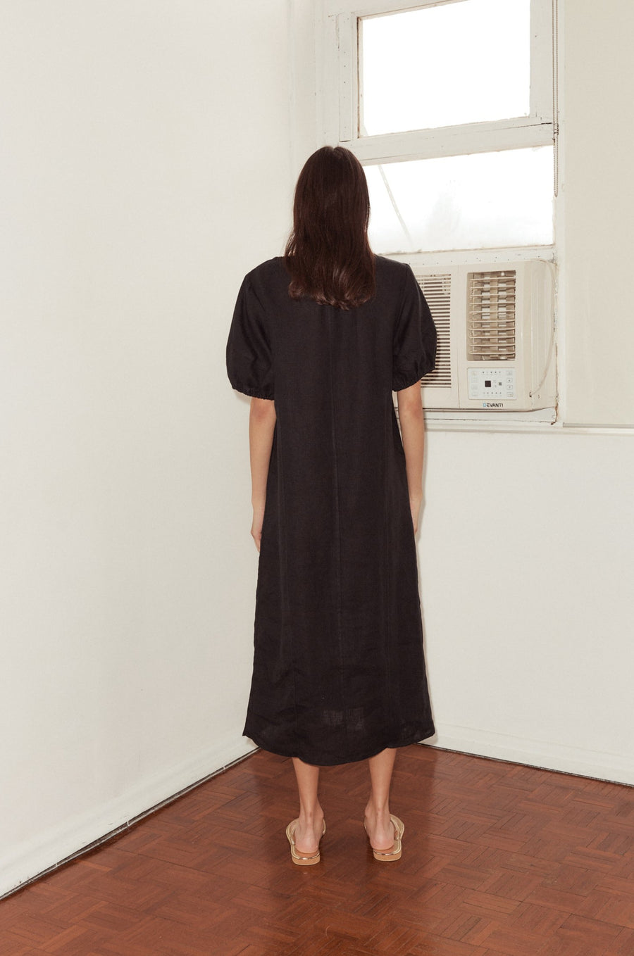 back shot of female model wearing the Curved Seam Midi Dress by Deiji Studios in black linen. The mid length dress features a bloussant sleeve with a soft elastic cuff and curated seam lines