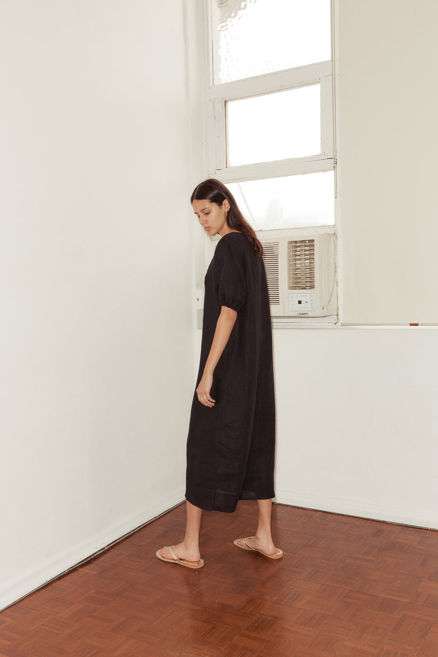 side shot of female model wearing the Curved Seam Midi Dress by Deiji Studios in black linen. The mid length dress features a bloussant sleeve with a soft elastic cuff and curated seam lines