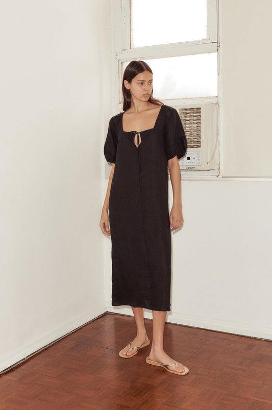 mid shot of female model wearing the Curved Seam Midi Dress by Deiji Studios in black linen. The mid length dress features an open square neckline with drawcord and bust key hole detail, bloussant sleeve with a soft elastic cuff and curated seam lines