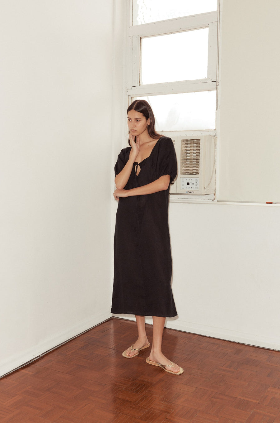 long shot of female model wearing the Curved Seam Midi Dress by Deiji Studios in black linen. The mid length dress features an open square neckline with drawcord and bust key hole detail, bloussant sleeve with a soft elastic cuff and curated seam lines
