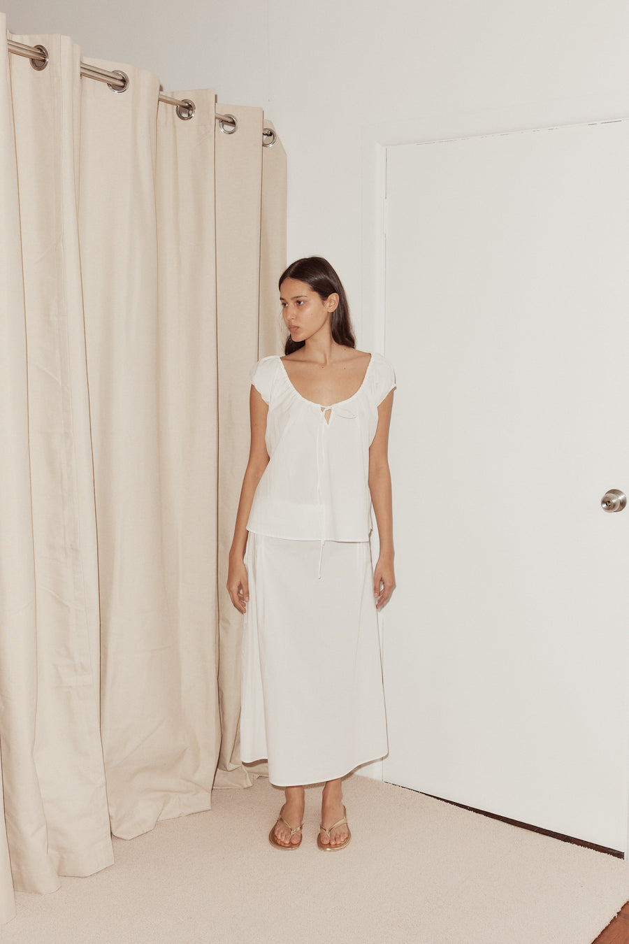 mid shot female model wearing one panel top by Deiji Studios in white. Elevated everyday top with capped sleeves, drawcord neckline with keyhole detail and a relaxed fit. Styled with pintuck skirt in white, relaxed and mid length.