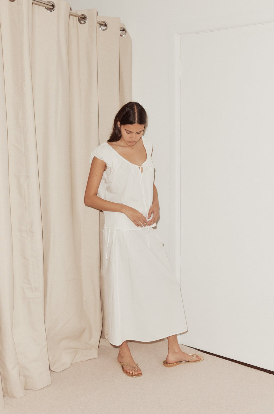 Side shot of female model wearing the Pintuck Skirt in white styled with the One Panel Top in white, standing on beige plush carpet in front of a beige curtain and white wall.