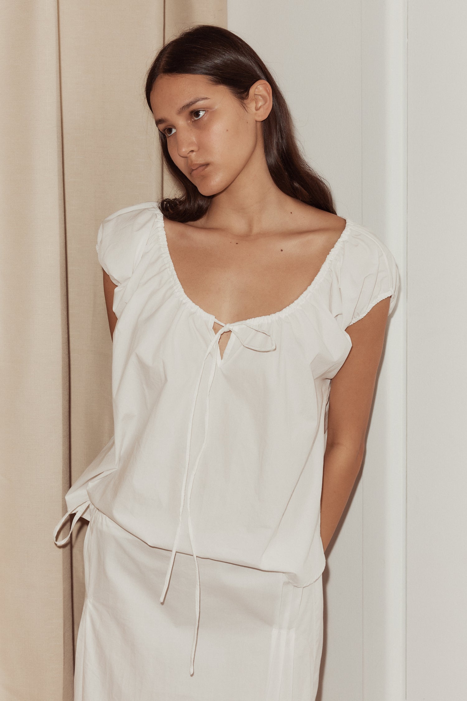 close up shot female model wearing one panel top by Deiji Studios in white. Elevated everyday top with capped sleeves features an adjustable gathered neckline with bow and keyhole detail. Styled with pintuck skirt in white.