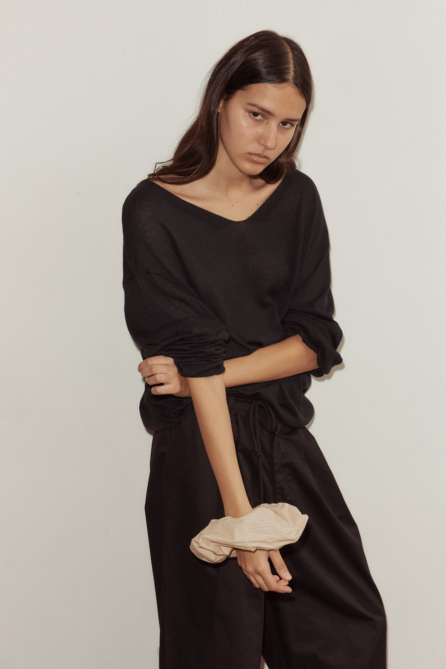 female model wears Deiji Studios loose long sleeve knitted top in black, rolled up to the elbows with scrunchie styled on wrist. A loose, open weave top with a soft v neck and elongated sleeves. Styled with the Deiji Studios Cotton Pant in black.