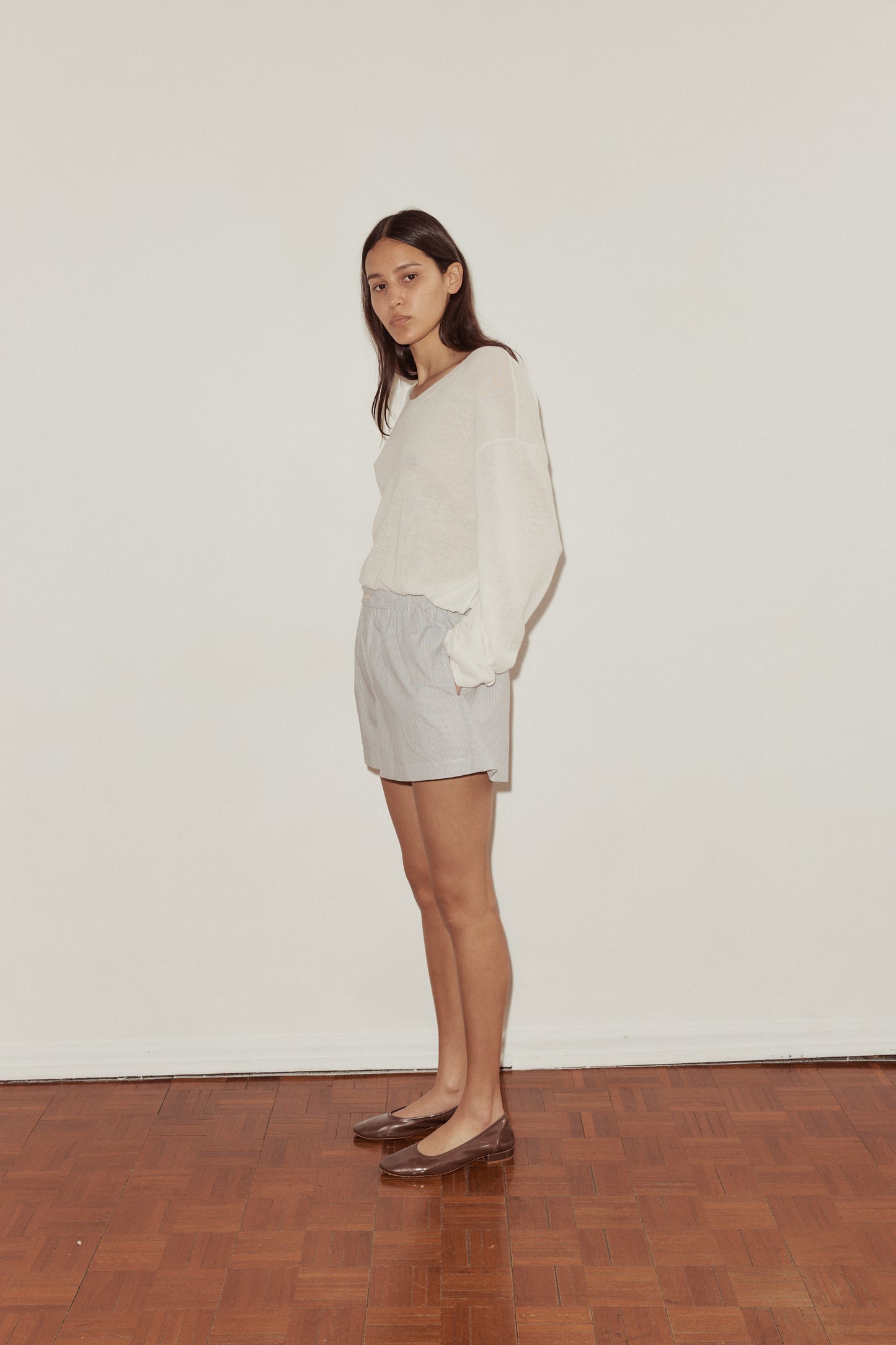 Side shot of female model wearing the Loose Long Sleeve Knitted Top by Deiji Studios in white, elongated sleeve folds over wrist of hand in Boxer short pocket.