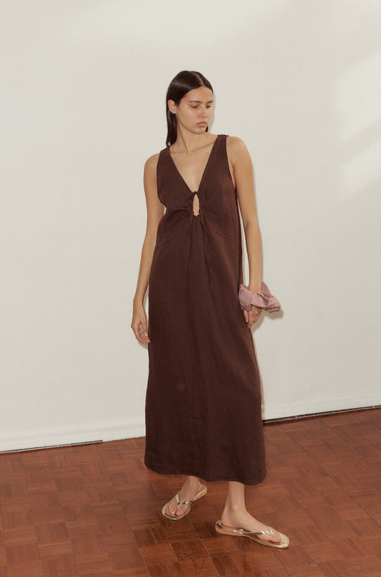 female model wears the Keyhole Dress by Deiji Studios in burgundy linen. Features a full length, flattering v neckline and drawcord keyhole detail at bust. Styled with Deiji Studios scrunchie in Lavender worn on wrist.