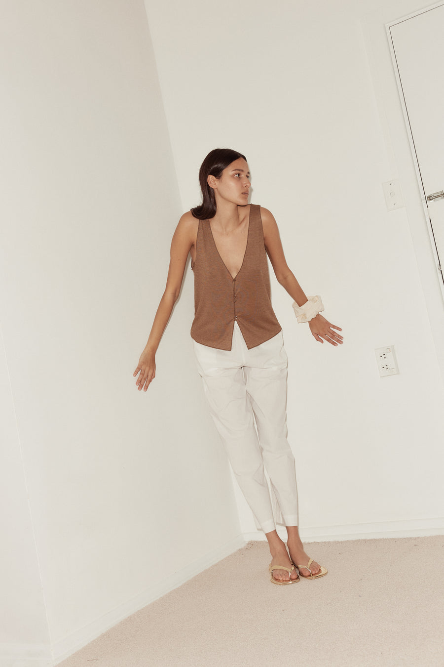 long shot of female model wearing the button up knit tank in coffee, features 4 buttons at front for adjustable opening and a deep v neckline. Styled with off white scrunchie worn on wrist and white ease trousers.