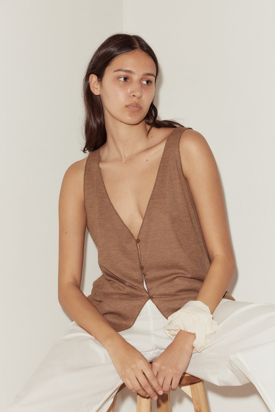 close up shot of female model wearing the button up knit tank in coffee, features 4 buttons at front for adjustable opening and a deep v neckline. Styled with off white scrunchie worn on wrist and white ease trousers.