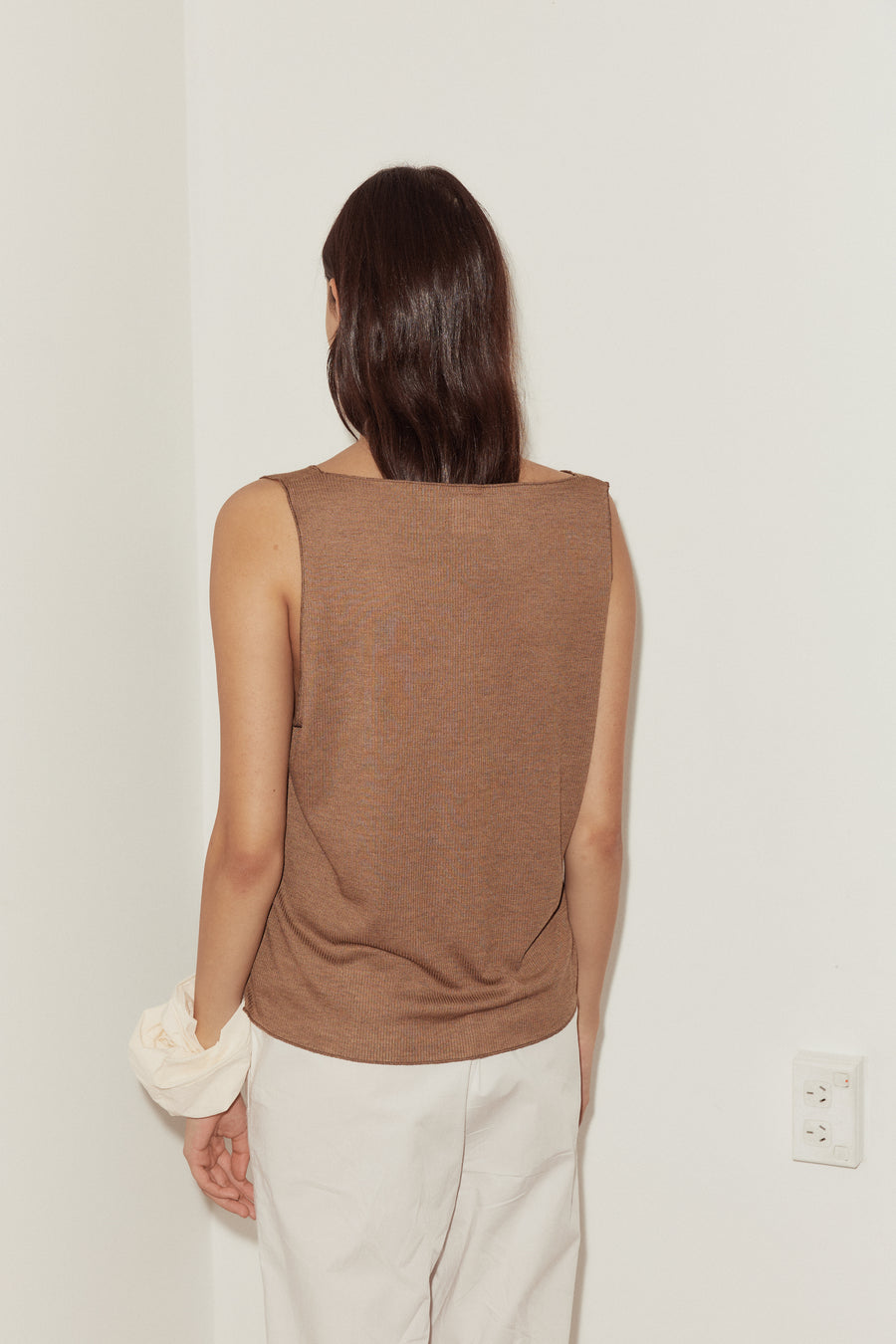 back shot of female model wearing the button up knit tank in coffee, features a minimal back with wide neckline. Styled with off white scrunchie worn on wrist and white ease trousers.