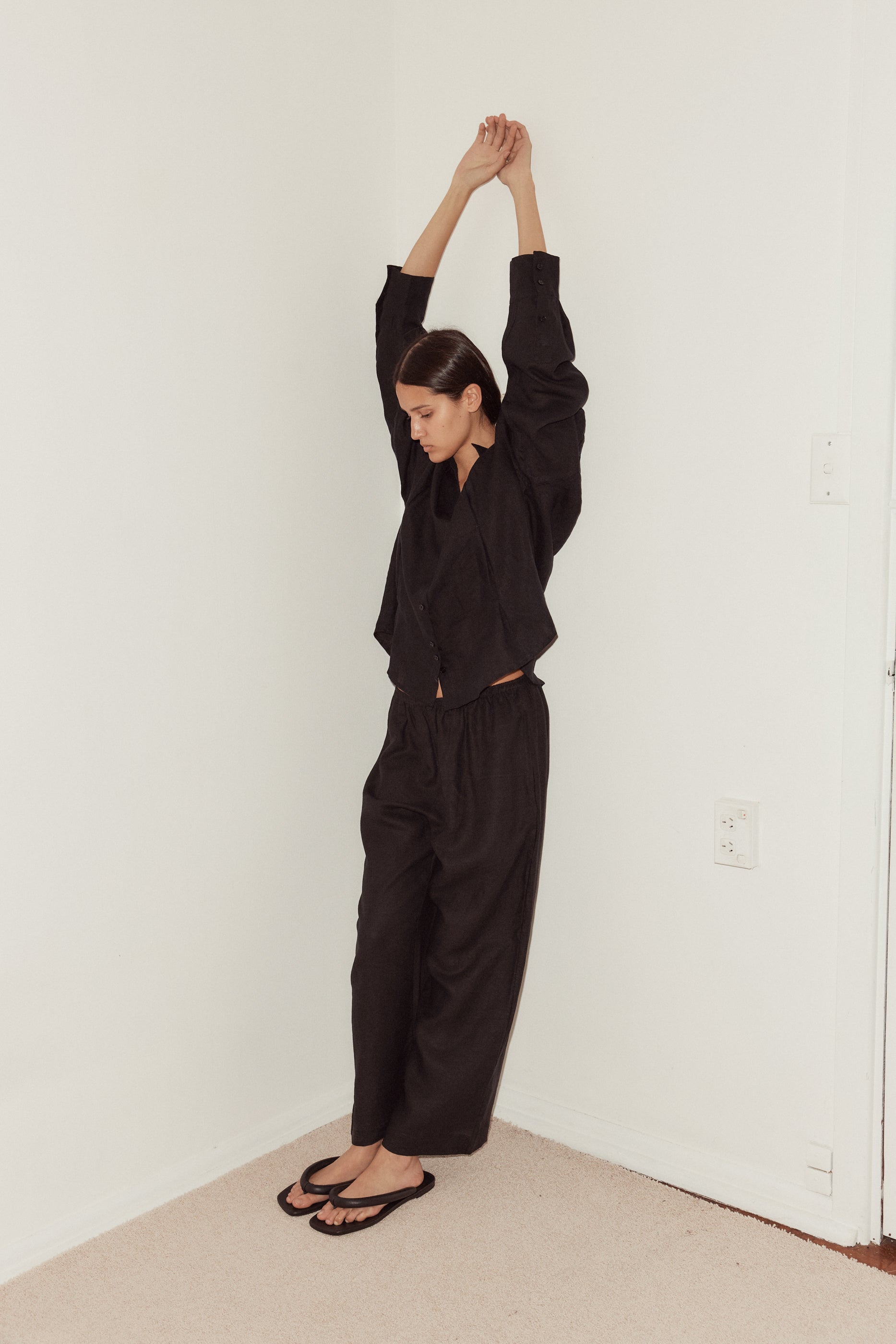 Female model standing in corner of white walls wearing the Tack Set by Deiji Studios in black, styled with off white scrunchie worn in hair. Tack Set pairs back with relaxed full length pant featuring soft elastic waist.