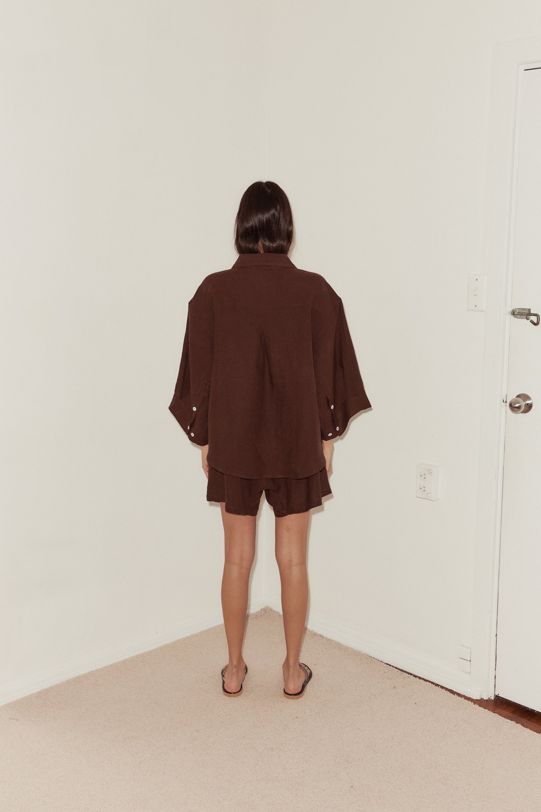 mid shot of female model with back to the camera wearing a burgundy Deiji Studios linen set, features an oversized shirt with pleat at back, button cuffs and relaxed shorts