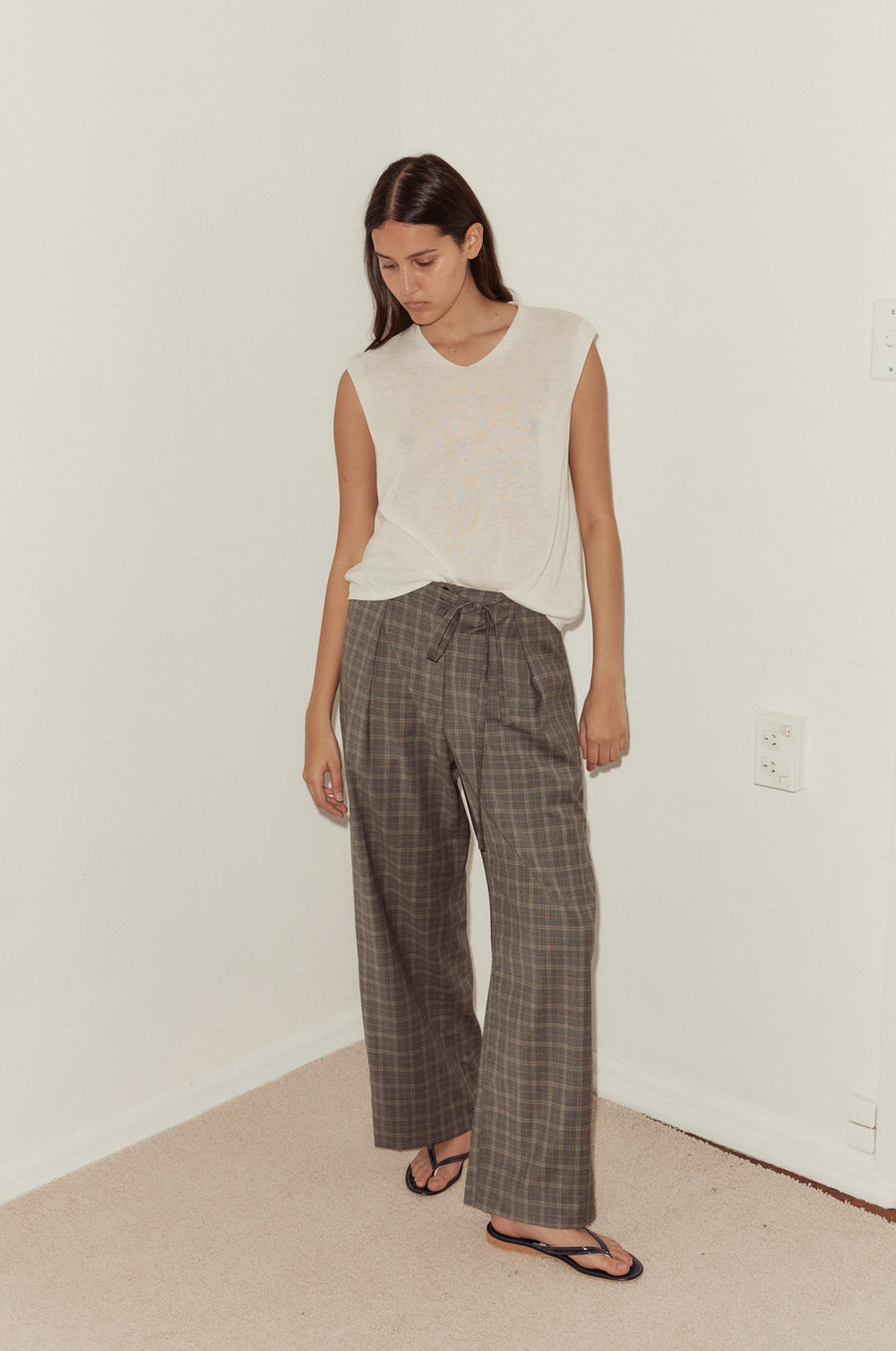 Mid shot of female model wearing the Tailored Pants in Everyday Check styled with the Loose Knitted Vest in white. Pant features relaxed leg, tailored pleat detail, button closure and self fabric tie at waist.