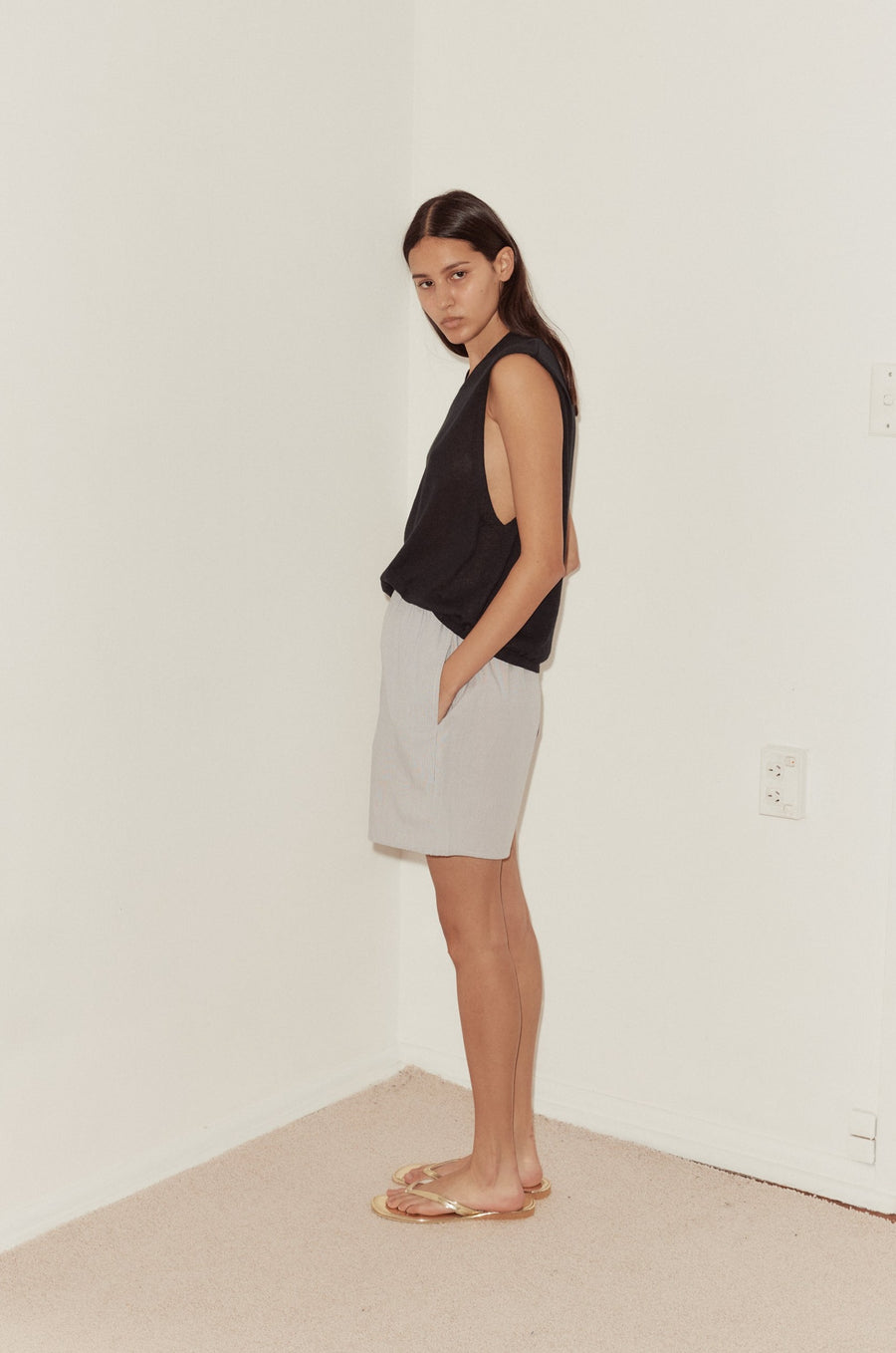 Side shot of female model with hand in shorts pocket wearing the Loose Knitted Vest by Deiji Studios in black. A lightweight knit vest with a soft high v neckline. Styled with the Mid Short by Deiji Studios in Dream stripe
