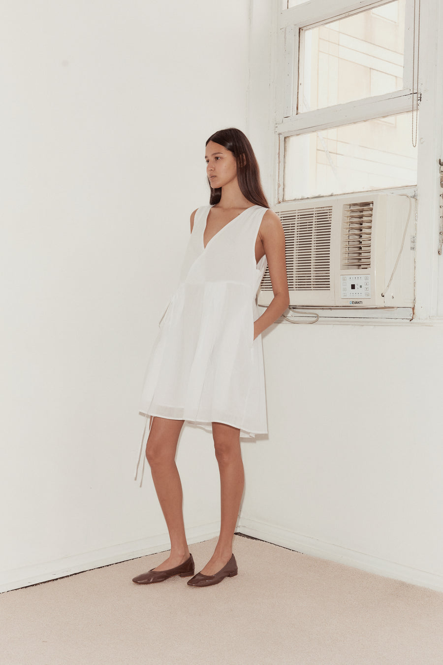 Mid shot of female model standing against a wall with a vintage airconditioning unit wearing the Wrap Tie Dress in white by Deiji Studios. A relaxed airy mini dress with cross front v neckline, waist tie and double lined skirt.